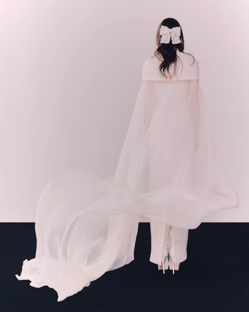 Back view of a woman in a white dress with a long train and a bow in her hair, standing against a white and black backdrop.