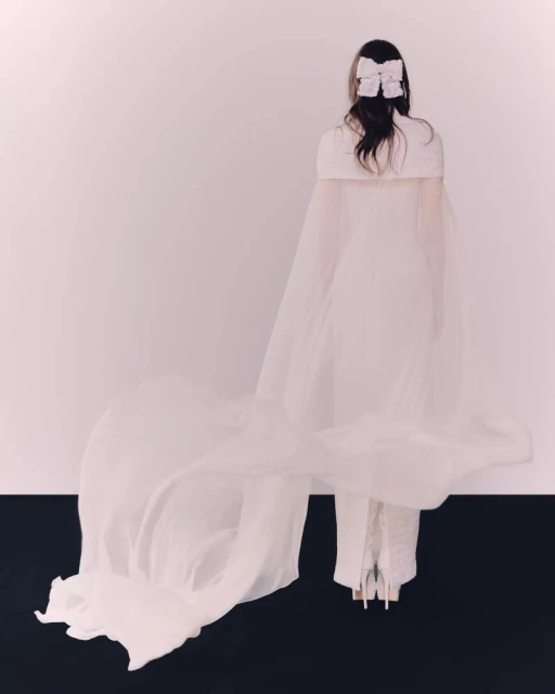 Back view of a woman in a white dress with a long train and a bow in her hair, standing against a white and black backdrop.