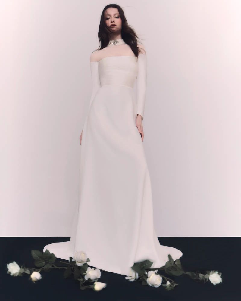 A woman in a long-sleeved, floor-length white gown with a sheer neckline poses in front of a gradient white backdrop with white roses.