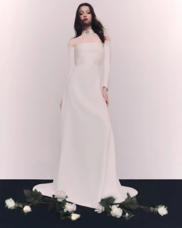A woman in a long-sleeved, floor-length white gown with a sheer neckline poses in front of a gradient white backdrop with white roses.
