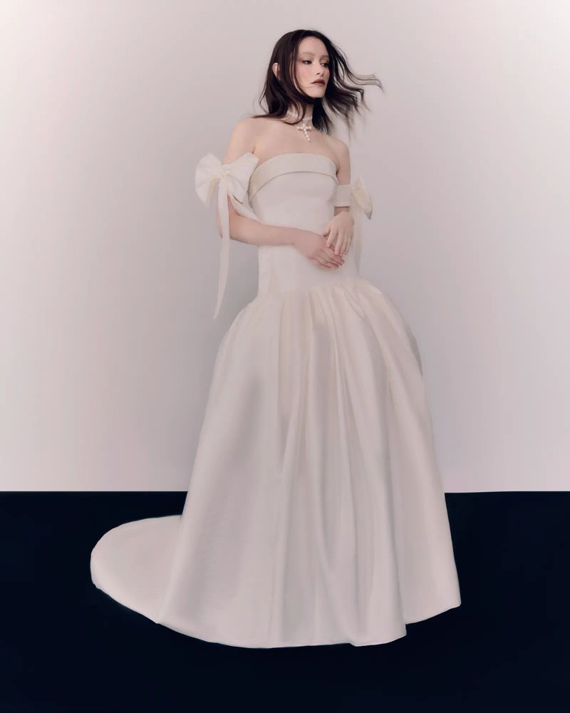 Elegant woman in a white strapless ballgown with bow details and a long train poses in a photo studio against a neutral backdrop.