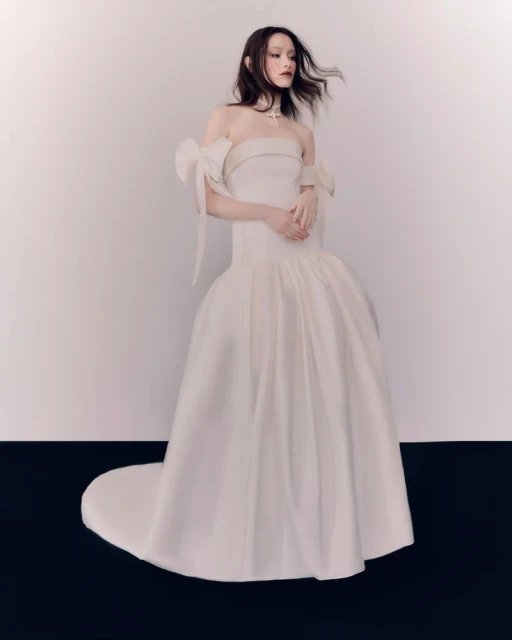 Elegant woman in a white strapless ballgown with bow details and a long train poses in a photo studio against a neutral backdrop.