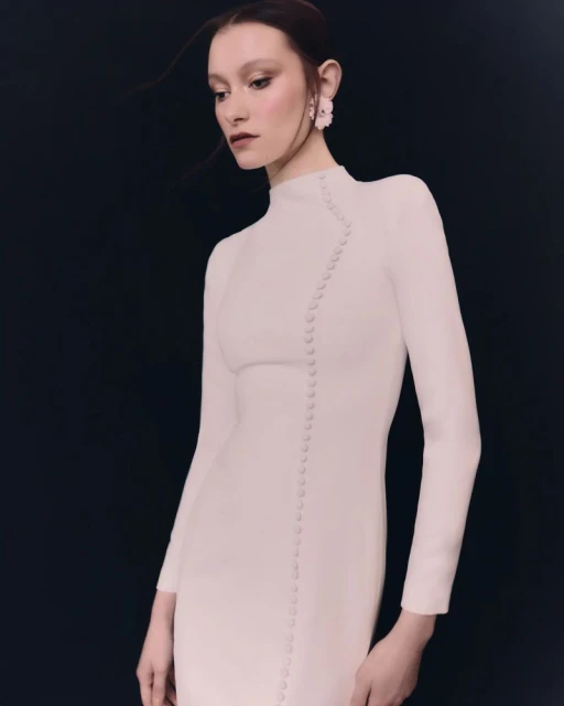 A woman poses in a fitted white dress with button accents against a dark background, showcasing fashion and elegant design.