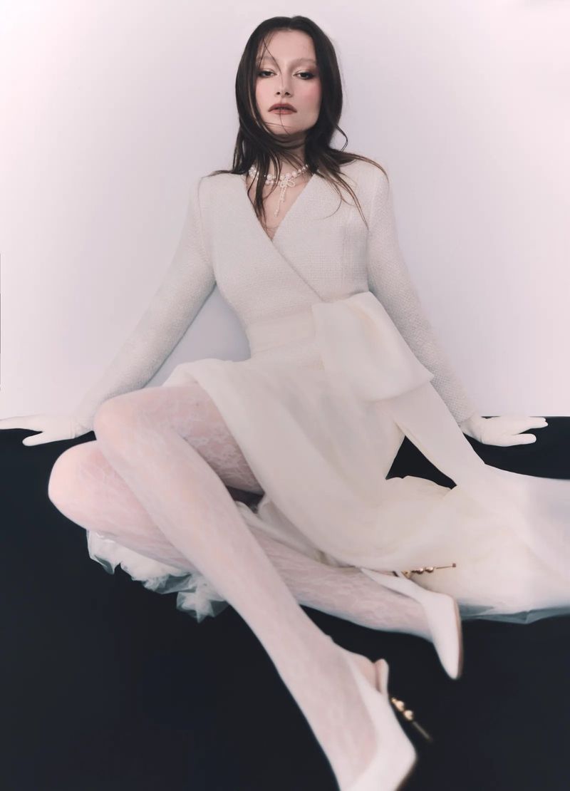 Woman poses seated wearing a white dress with sheer skirt and lace tights against a black and white backdrop in a studio shot.