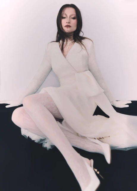Woman poses seated wearing a white dress with sheer skirt and lace tights against a black and white backdrop in a studio shot.