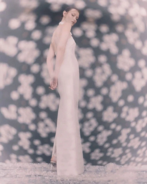 Woman in a white dress poses against a blue floral backdrop in a fashion-style shot, featuring soft focus and an elegant aesthetic.