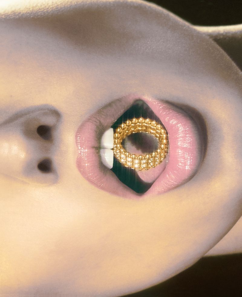 Woman's face in close-up featuring a golden ring held in the mouth between the lips and teeth, creating a striking, surreal aesthetic.