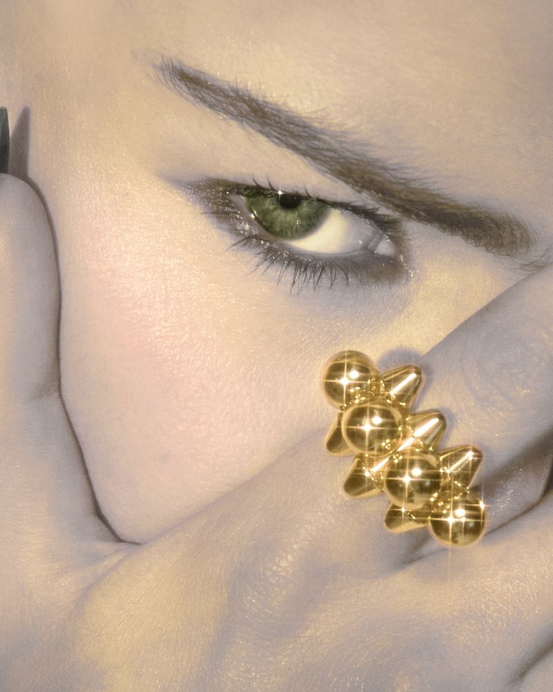 Close-up portrait of a person with an eye gazing towards the camera, wearing a gold ring on their finger with glossy makeup.