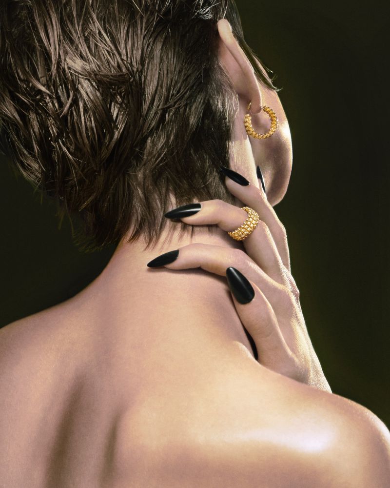 Close-up of a person's back and head, wearing gold earrings and a ring, with long black fingernails against a dark background.