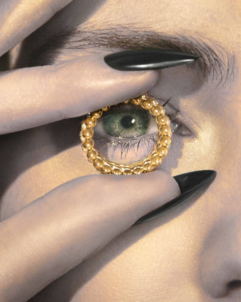 Close-up portrait of a woman's face with a gold beaded ring held up to her eye, emphasizing fashion and beauty with long, dark nails.