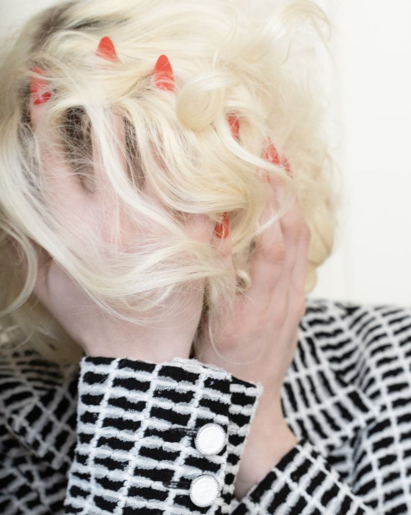 Woman with blonde hair covering face and wearing patterned jacket with visible buttons in a blurred indoor setting.