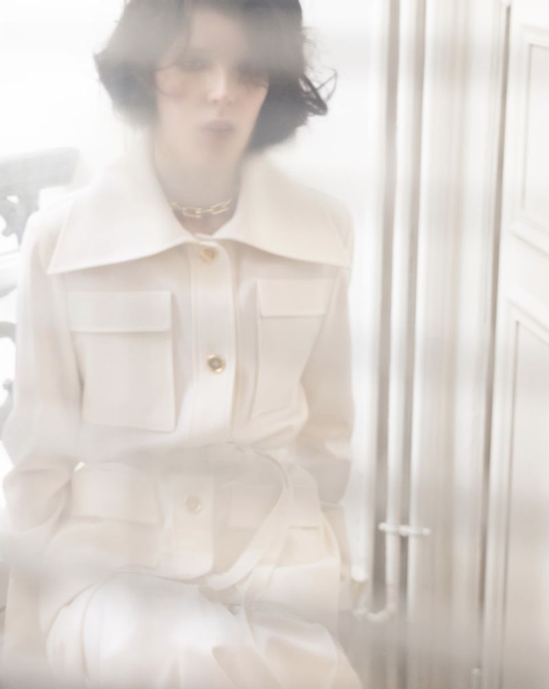 A blurred, dreamy fashion shot of a woman in a white outfit, featuring a tailored jacket and matching pants in a soft, diffused light.