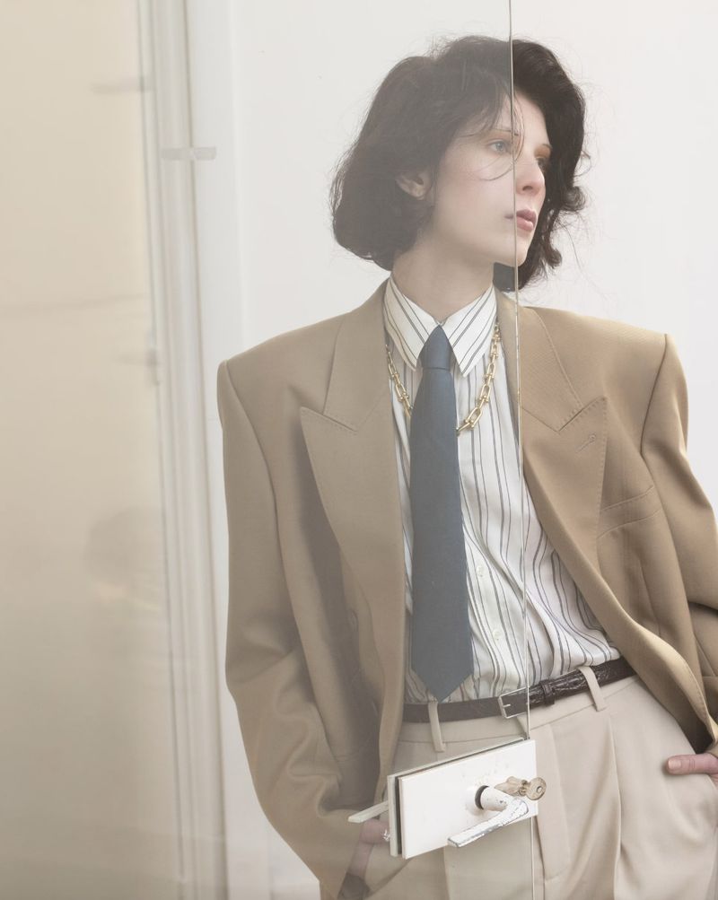 A woman with short dark hair and wearing a beige blazer, striped shirt, dark tie, and gold necklace is captured indoors.