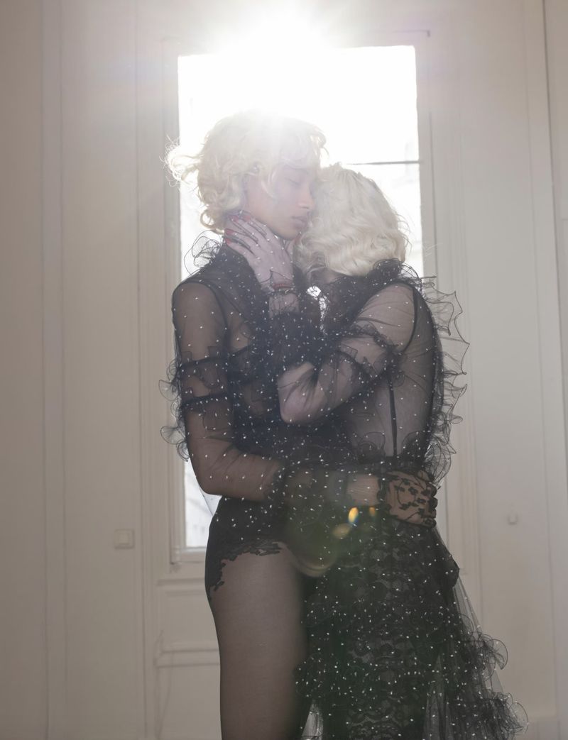 Two women embrace, wearing black lace dresses in a room with a bright window. Ethereal fashion shot with warm, diffused lighting.