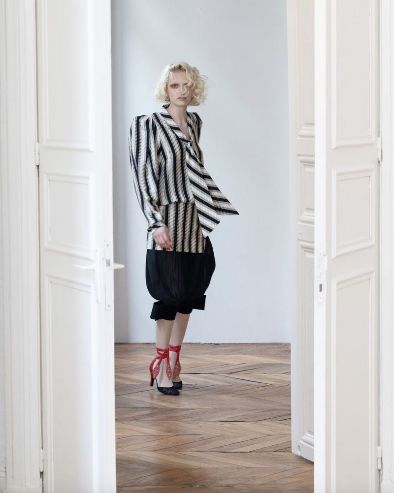 Fashionable blonde woman in striped jacket, pleated culottes, and red heeled shoes poses in doorway. Fashion editorial shot in bright white interior.