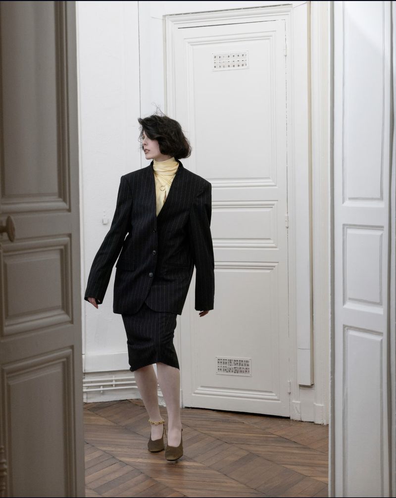 Woman in a dark pinstripe skirt suit walks through a doorway into a room with herringbone wood floors and white walls and door frames.