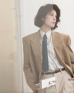 Model in oversized camel blazer wearing a striped shirt, tie, and carrying a structured white handbag