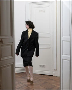 Model in a black pinstripe blazer and matching skirt with a yellow turtleneck walking indoors on hardwood parquet flooring.