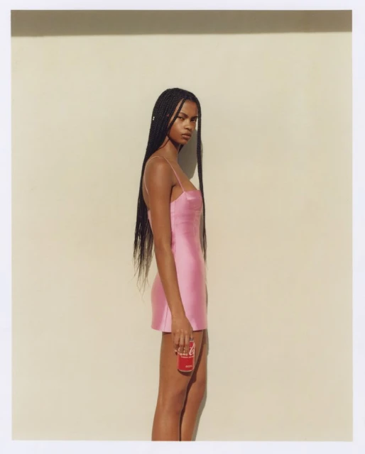 Profile shot of a young Black woman in a pink dress holding a Coca-Cola can against a light yellow wall, simple studio setting.