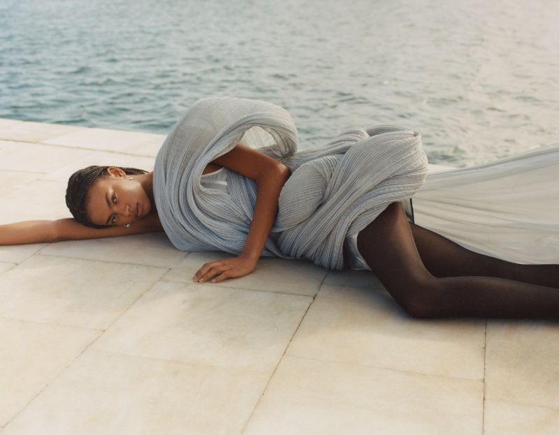 Fashion shot of a model in a draped grey gown, lying on tiled surface with a water background, capturing elegance and serenity.