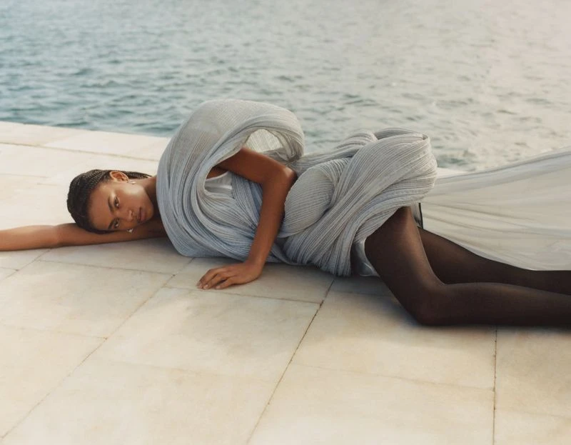 Fashion shot of a model in a draped grey gown, lying on tiled surface with a water background, capturing elegance and serenity.