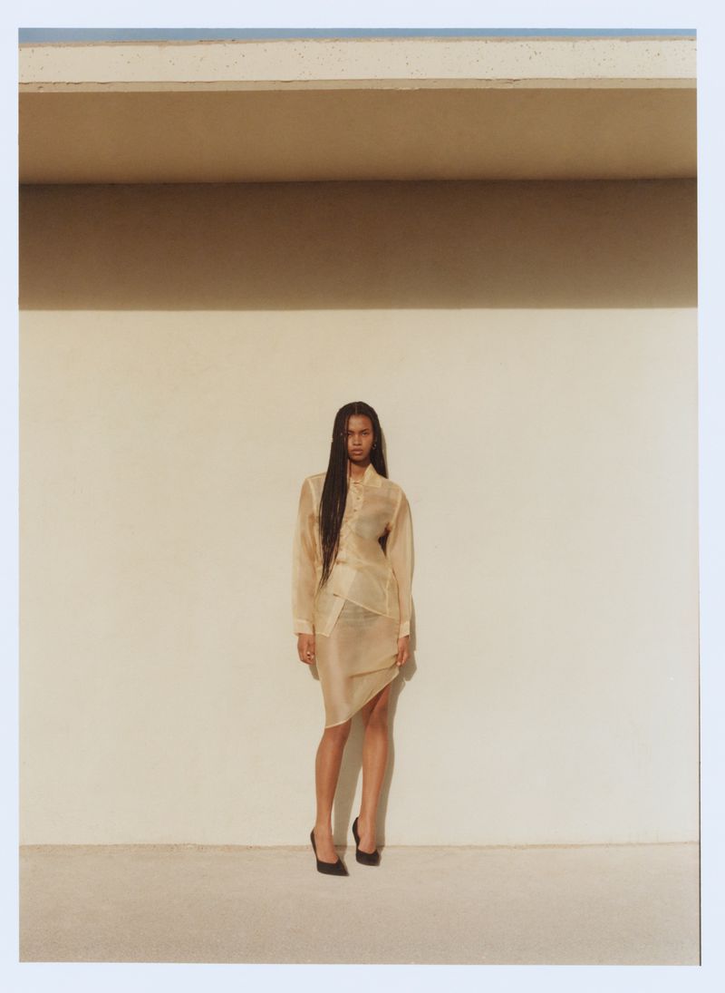 A woman poses in a beige skirt and blouse in front of a neutral-colored wall in an indoor studio setting with natural light.