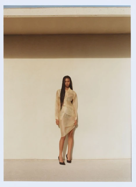 A woman poses in a beige skirt and blouse in front of a neutral-colored wall in an indoor studio setting with natural light.