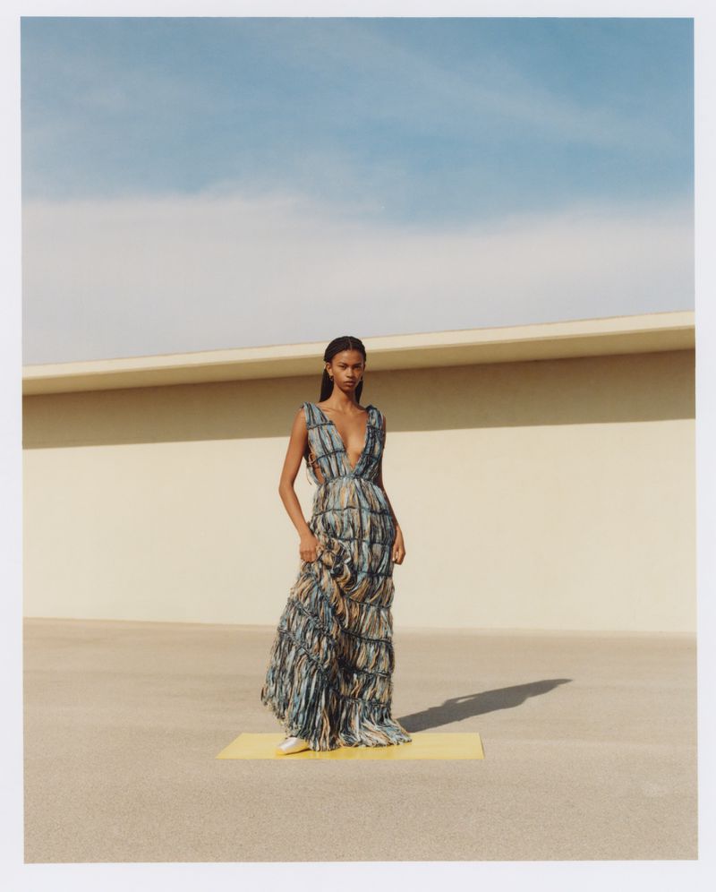 A single woman poses in a full-length print dress with a plunging neckline against a sunlit wall and blue sky in a fashion editorial.