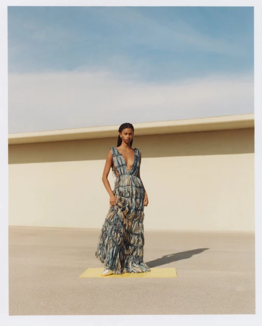 A single woman poses in a full-length print dress with a plunging neckline against a sunlit wall and blue sky in a fashion editorial.