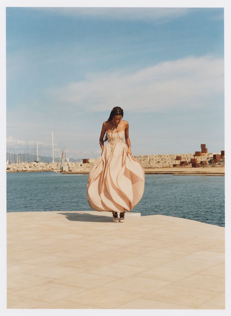 A solitary woman in a flowing gown stands on a tan platform by the sea, under a bright blue sky, with a harbor visible in the background.