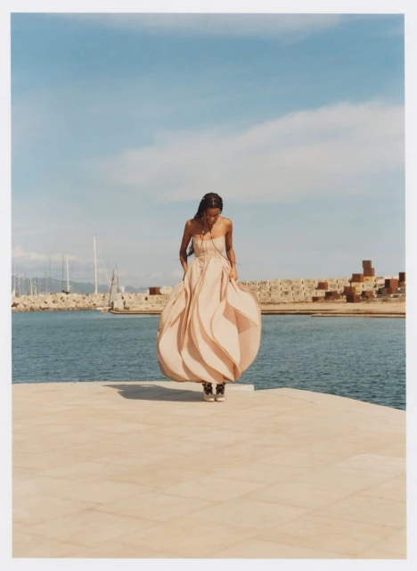 A solitary woman in a flowing gown stands on a tan platform by the sea, under a bright blue sky, with a harbor visible in the background.