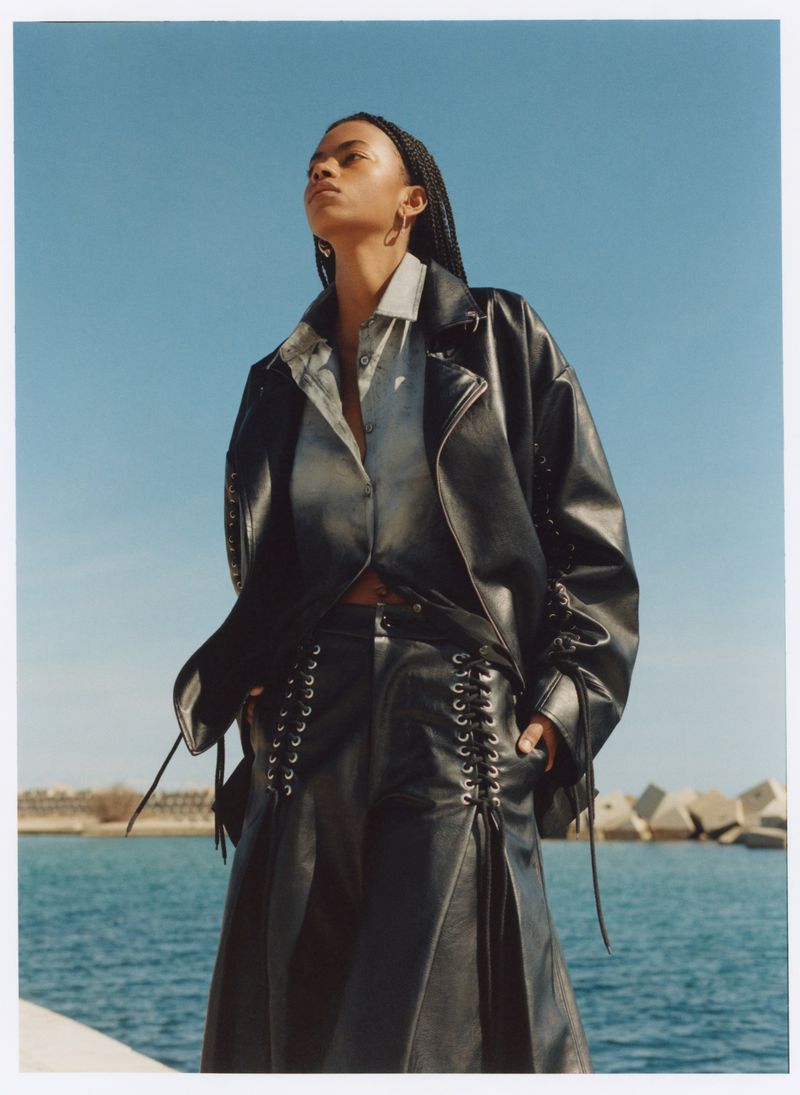 Model in black leather jacket and matching pants standing outdoors against an ocean and clear sky background.