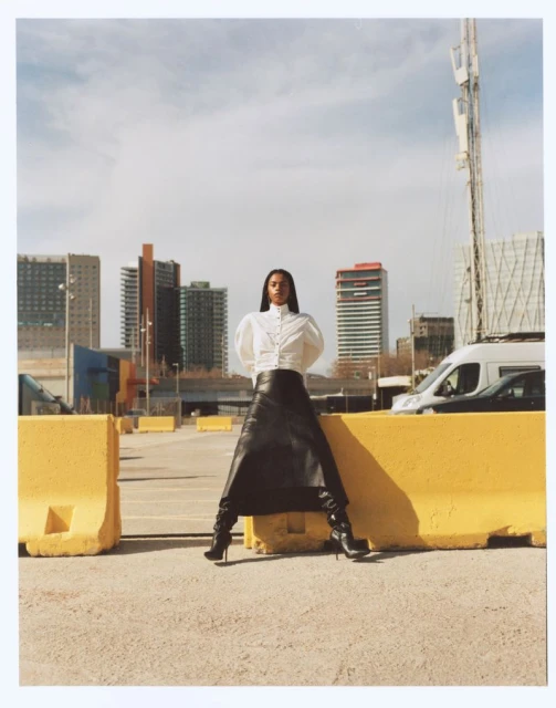 Fashion shot of a woman in a white blouse and long black skirt, posing outdoors against yellow barriers and a city skyline backdrop.