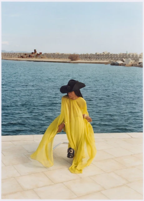 Woman in a yellow dress and black hat poses on a tiled surface with the sea and coastal buildings in the background under a bright sky.