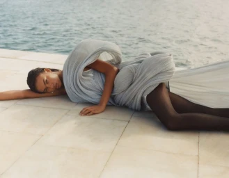 Model lying down on a tiled terrace wearing a draped grey dress with water visible in the background