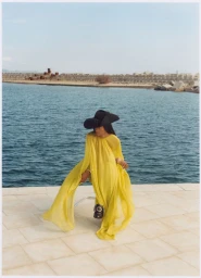Model in yellow dress and wide black hat seated by the water for Schön Magazine editorial Nautilust 080.