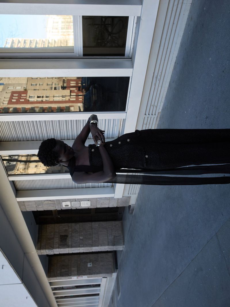 Adult woman poses wearing a black jumpsuit with silver bracelets against a modern building with large windows in an urban area during daytime.