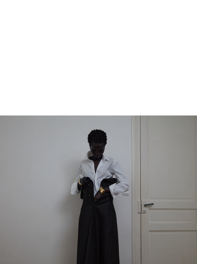 Dark-skinned woman zips up long black skirt, wearing white blouse and gold bracelets in a minimalist interior with white wall and door.