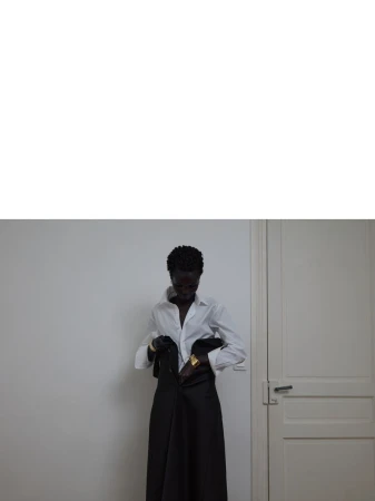 Dark-skinned woman zips up long black skirt, wearing white blouse and gold bracelets in a minimalist interior with white wall and door.