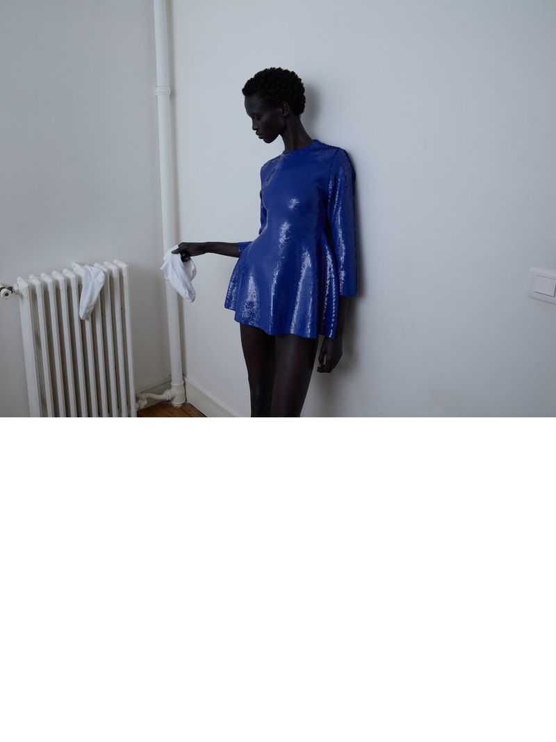 Elegant dark-skinned woman in a shiny blue mini-dress leans against a white wall, holding a white cloth, next to a radiator.