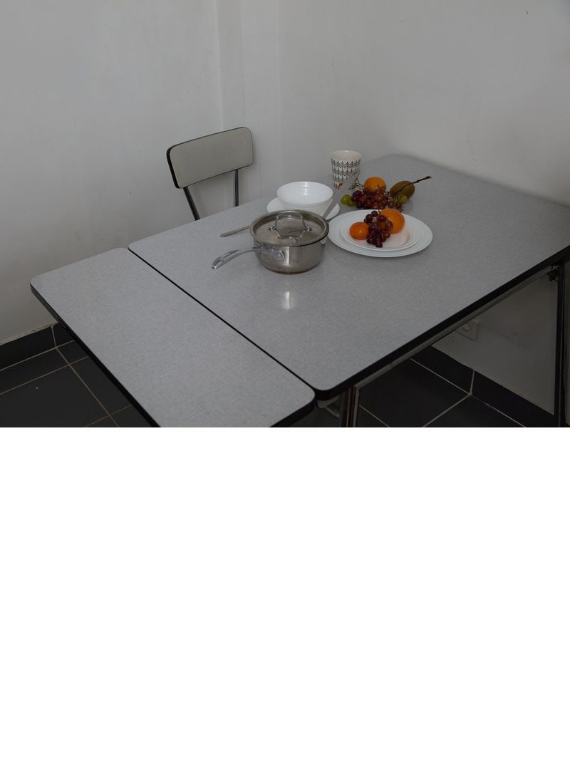 Still life featuring a pot with lid, fruit, tableware, and a chair, arranged on a gray folding table in an indoor setting against a white wall.