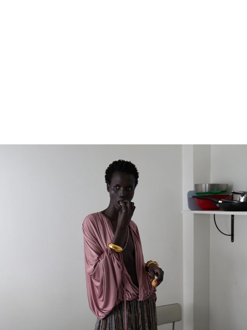 Dark-skinned adult woman in a pink top and patterned skirt poses in a white room with kitchenware on a shelf in the background.