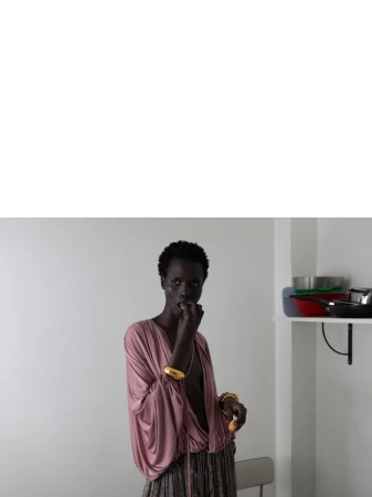 Dark-skinned adult woman in a pink top and patterned skirt poses in a white room with kitchenware on a shelf in the background.