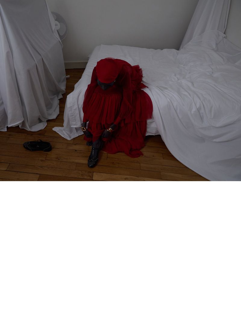 Person shrouded in red fabric sits on a bed with white linens in a simple room, exuding a sense of mystery and concealment.