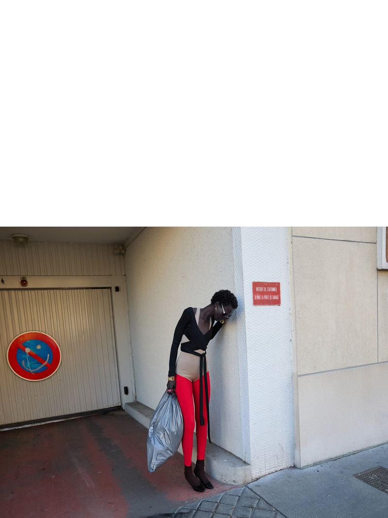 A slender woman in red leggings leans against a wall in an urban setting, holding a trash bag, next to a no-parking sign.