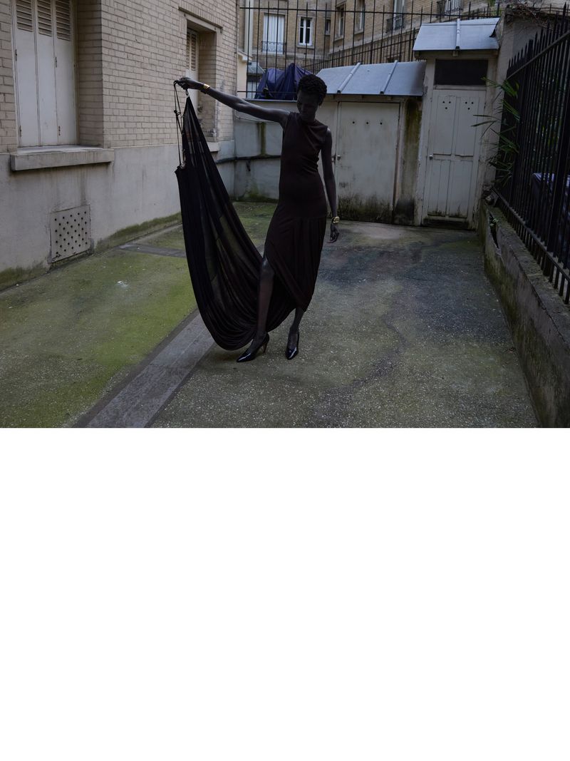 Dark-skinned woman in draped black dress poses in urban alley holding hammock, wearing heels, metal bracelet, outdoors, daytime.