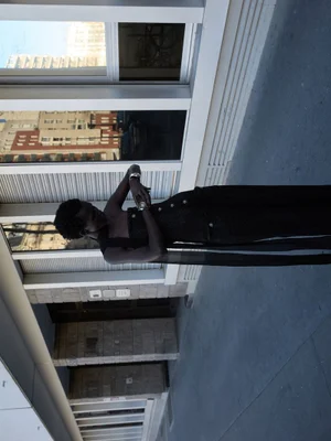 Model Amar Akway in a black textured outfit standing outdoors against an architectural window and stone wall background.