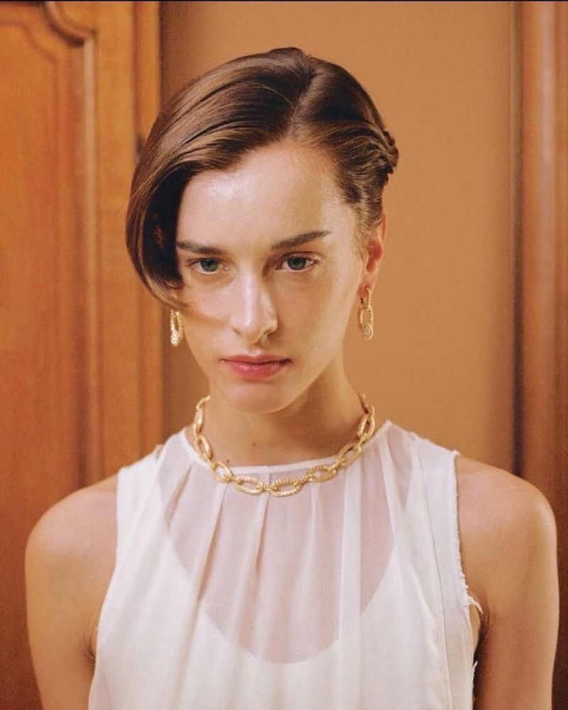 Portrait of a woman wearing a white sleeveless top and gold chain necklace against an indoor wood-paneled wall