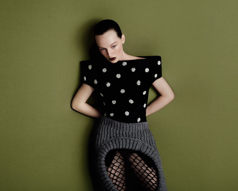 A woman with dark hair and makeup poses in a studio. She is wearing a black top with white polka dots and a grey knit skirt.