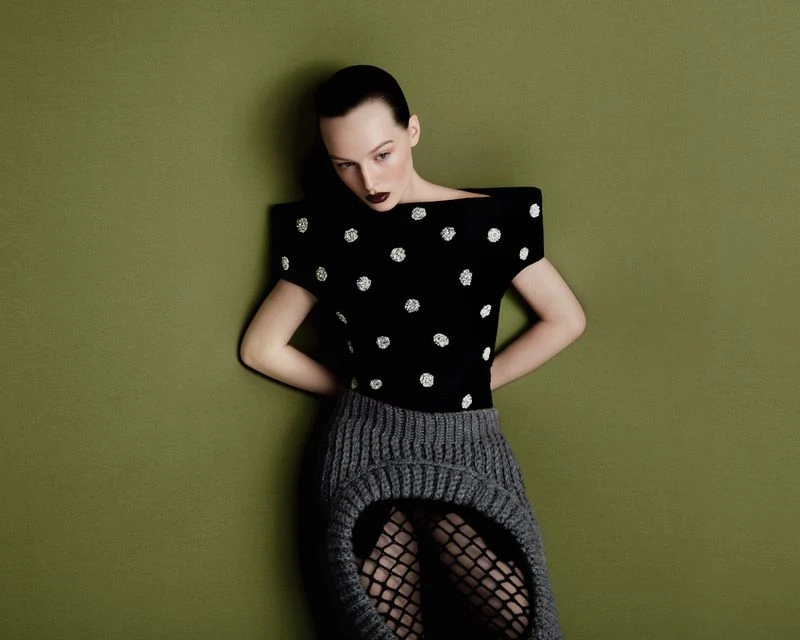 A woman with dark hair and makeup poses in a studio. She is wearing a black top with white polka dots and a grey knit skirt.